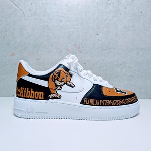 Könnte beinhalten: Weißer, schwarzer und orangefarbener Sportschuh mit einem Tiger-Motiv und dem Text "Florida International University". Der Schuh hat eine weiße Sohle und Schnürsenkel. Das Wort "McKibbon" ist ebenfalls auf der Seite zu sehen.