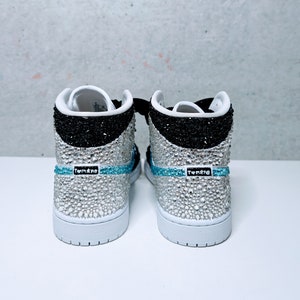 May include: A pair of white high-top sneakers with black and silver accents. The sneakers are covered in rhinestones and have a blue stripe with the word "Tomkre" on them.