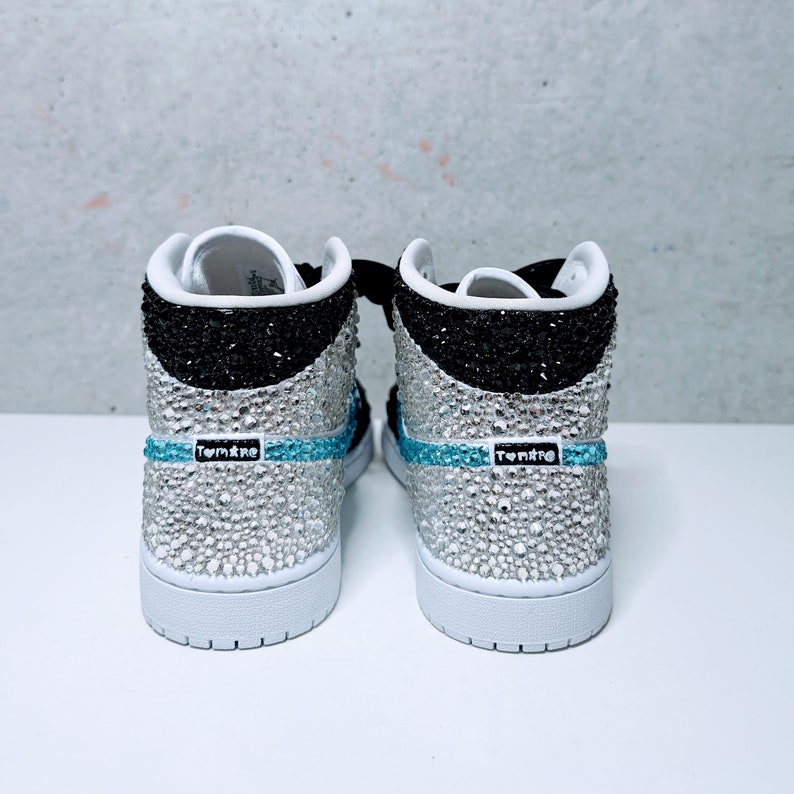 May include: A pair of white high-top sneakers with black and silver accents. The sneakers are covered in rhinestones and have a blue stripe with the word "Tomkre" on them.