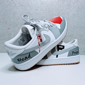 May include: A pair of white low-top sneakers with gray and red accents. The left shoe has the word "Head" written on the side, and the right shoe has the word "Friar" written on the side.