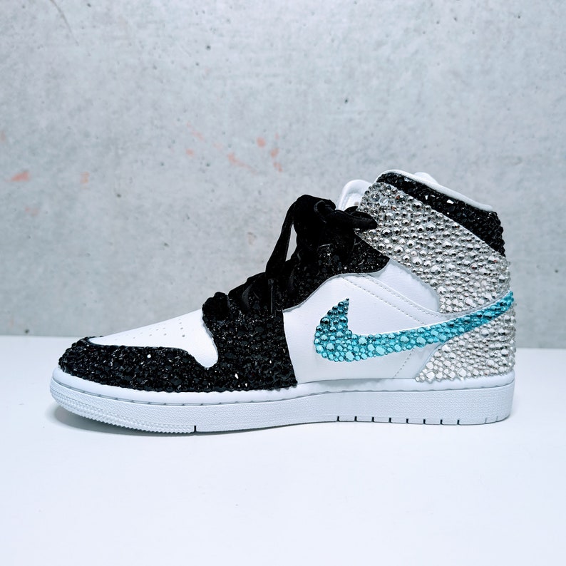 May include: White, black, and blue high-top sneakers with a crystal-encrusted design. The sneakers have a white sole, a black leather upper, and a blue swoosh logo.