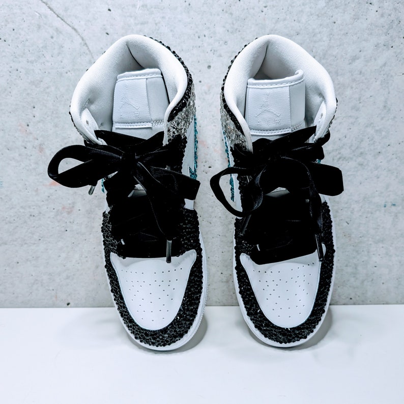 May include: A pair of white high-top sneakers with black trim and a sparkly design. The sneakers have black laces and a black ribbon tied around the ankles.