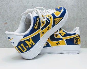 Puede incluir: Un par de zapatillas blancas con detalles pintados en azul y amarillo. Las zapatillas tienen las palabras "MICHIGAN" y "U.M." pintadas en ellas. Las zapatillas también tienen un logotipo swoosh amarillo y una pequeña mascota de glotón pintada en ellas.