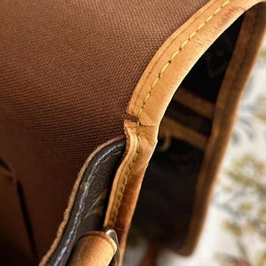 May include: Close-up of a brown canvas bag with tan leather trim. The bag features yellow stitching along the edges and a patterned interior. The leather has a slight sheen.
