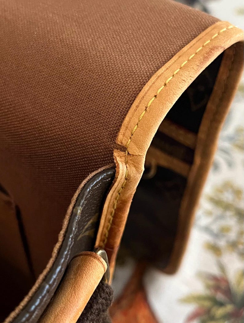 May include: Close-up of a brown canvas bag with tan leather trim. The bag features yellow stitching along the edges and a patterned interior. The leather has a slight sheen.