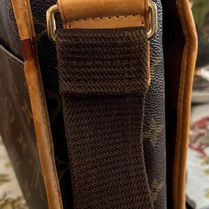 May include: Close-up of a brown messenger bag with a canvas strap and leather trim. The bag features a repeating pattern and gold-colored hardware. The strap is a dark brown color.