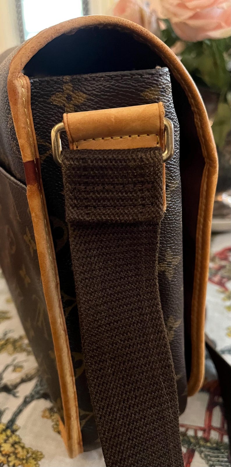 May include: Close-up of a brown messenger bag with a canvas strap and leather trim. The bag features a repeating pattern and gold-colored hardware. The strap is a dark brown color.