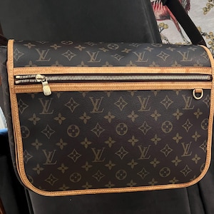 May include: Brown messenger bag with a repeating monogram pattern. The bag features a gold zipper, a tan leather trim, and a gold-colored metal ring. The bag has a shoulder strap.
