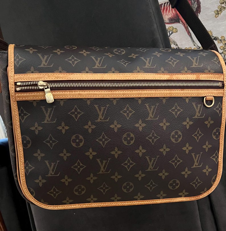 May include: Brown messenger bag with a repeating monogram pattern. The bag features a gold zipper, a tan leather trim, and a gold-colored metal ring. The bag has a shoulder strap.