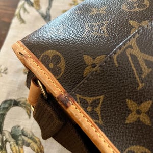 May include: Close-up of a brown and tan handbag with a repeating logo pattern. The bag features a brown strap and tan leather trim with visible stitching. The bag's corner shows wear and tear.