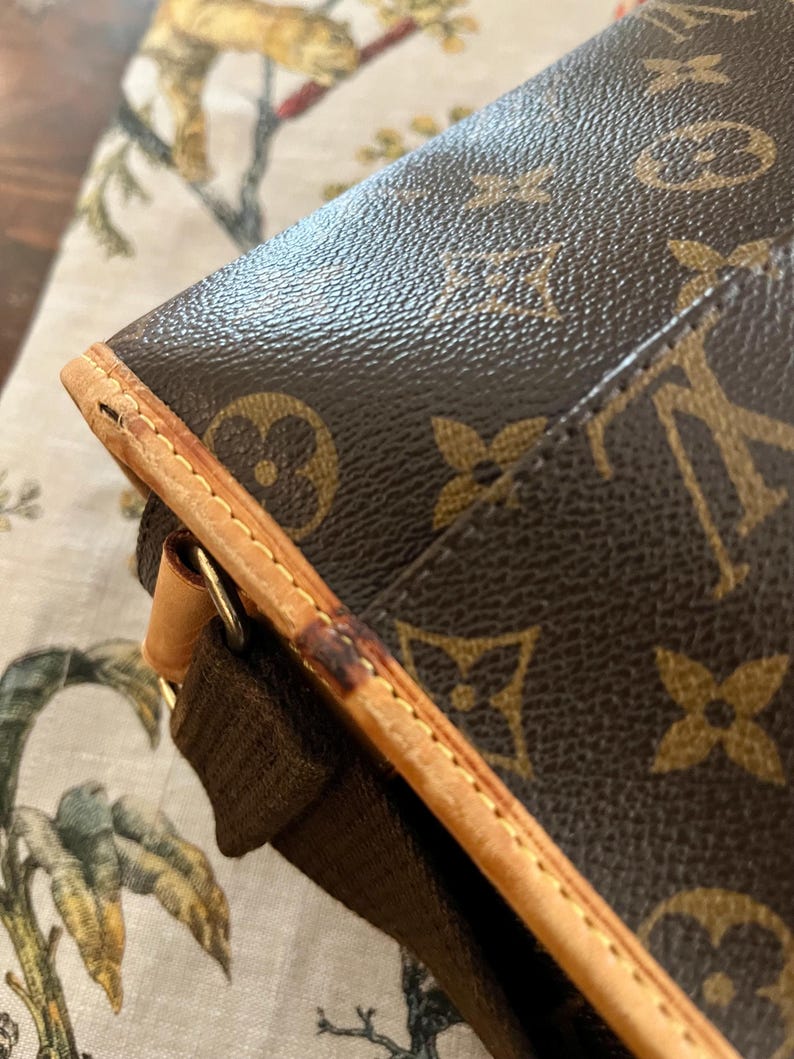 May include: Close-up of a brown and tan handbag with a repeating logo pattern. The bag features a brown strap and tan leather trim with visible stitching. The bag's corner shows wear and tear.