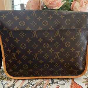 May include: Brown messenger bag with a repeating pattern of interlocking letters and floral designs. The bag has a light brown trim and a black shoulder strap. The bag is sitting on a patterned surface.