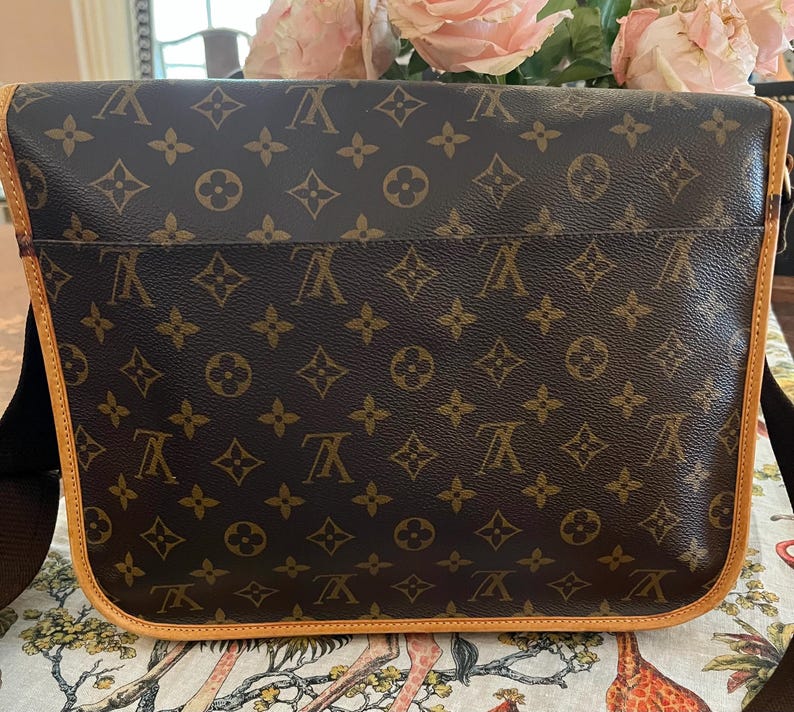 May include: Brown messenger bag with a repeating pattern of interlocking letters and floral designs. The bag has a light brown trim and a black shoulder strap. The bag is sitting on a patterned surface.