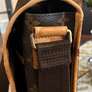 May include: Close-up of a brown and tan shoulder bag. The bag features a brown canvas strap and a leather trim. The bag's exterior has a repeating pattern of brown floral designs. The bag's hardware is gold-colored.