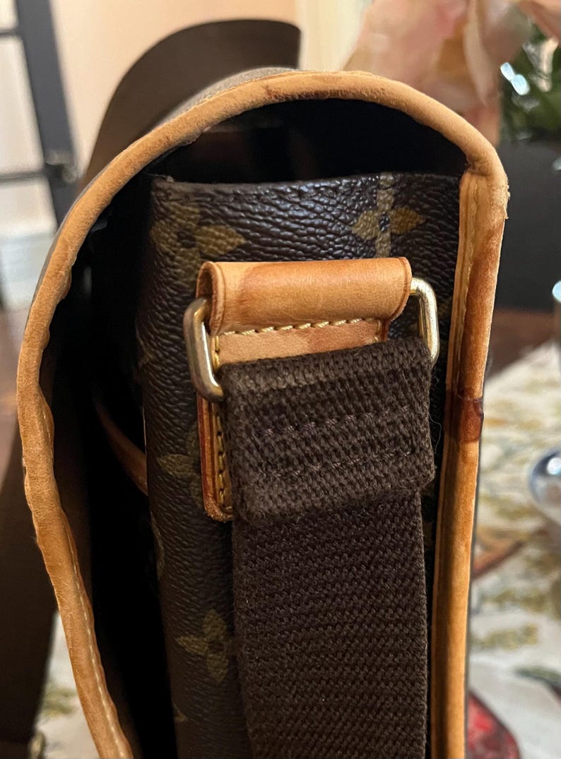 May include: Close-up of a brown and tan shoulder bag. The bag features a brown canvas strap and a leather trim. The bag's exterior has a repeating pattern of brown floral designs. The bag's hardware is gold-colored.