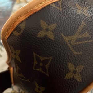 May include: Close-up of a brown and tan handbag. The bag features a repeating pattern of brown floral shapes on a dark brown background. The edges are trimmed with tan leather and stitched with tan thread.