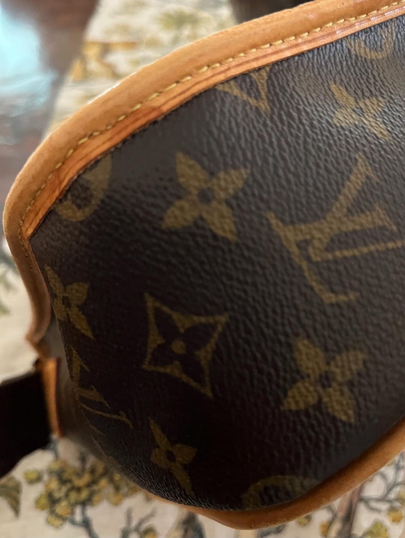 May include: Close-up of a brown and tan handbag. The bag features a repeating pattern of brown floral shapes on a dark brown background. The edges are trimmed with tan leather and stitched with tan thread.