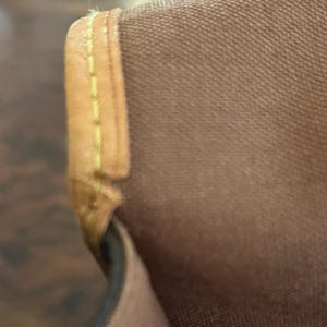 May include: Close-up of a brown fabric with a tan leather trim. The leather trim is stitched with yellow thread. The fabric appears to be a durable material, possibly canvas or a similar textile. The image focuses on the corner of a bag or similar item.