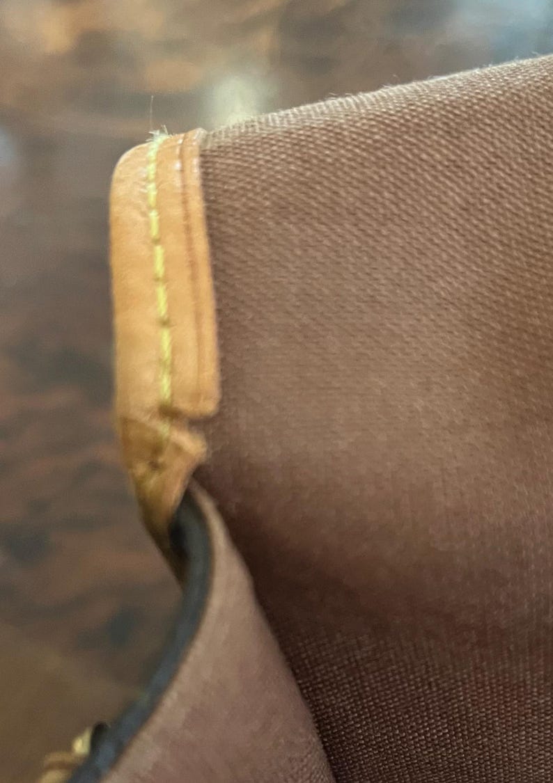 May include: Close-up of a brown fabric with a tan leather trim. The leather trim is stitched with yellow thread. The fabric appears to be a durable material, possibly canvas or a similar textile. The image focuses on the corner of a bag or similar item.