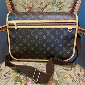May include: Brown messenger bag with a repeating monogram pattern. The bag features a zippered top, a brown adjustable shoulder strap, and tan trim. The bag is sitting on a floral patterned chair.
