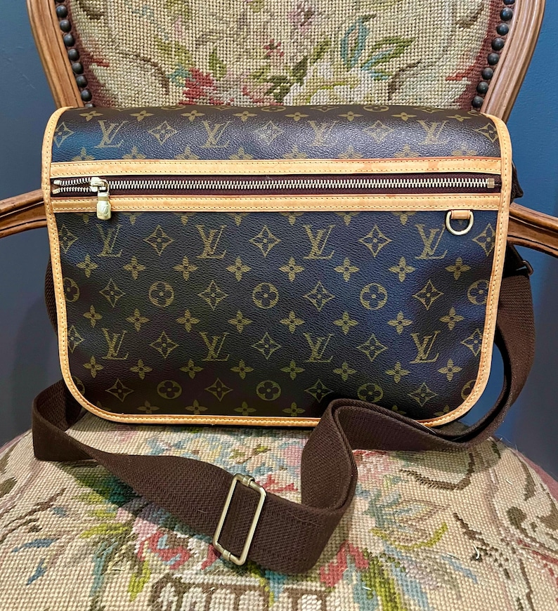 May include: Brown messenger bag with a repeating monogram pattern. The bag features a zippered top, a brown adjustable shoulder strap, and tan trim. The bag is sitting on a floral patterned chair.