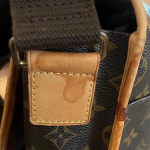 May include: Close-up of a brown and tan shoulder bag with a repeating logo pattern. The bag features a brown strap and tan leather accents. The leather tag shows a water stain. The bag's trim is a lighter tan color.