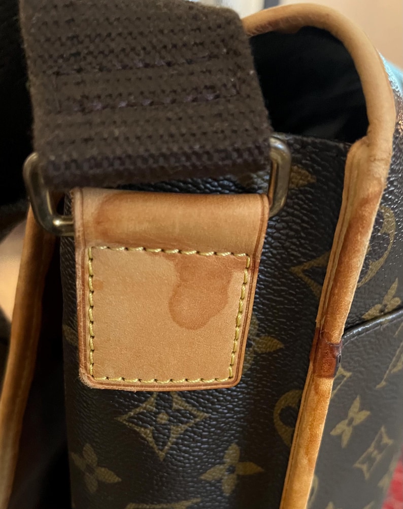 May include: Close-up of a brown and tan shoulder bag with a repeating logo pattern. The bag features a brown strap and tan leather accents. The leather tag shows a water stain. The bag's trim is a lighter tan color.