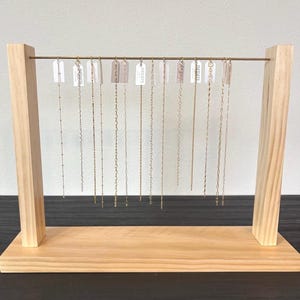 Op de afbeelding: Een sieradenstandaard van lichtgekleurd hout met een horizontale metalen stang. Twaalf dunne goudkleurige kettingen hangen aan de stang, elk bevestigd aan een klein wit rechthoekig label met het woord INFINITE in zwarte letters op de labels.