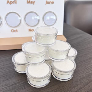 以下が含まれることがあります： 細かい白い粉が入った、小さく透明な丸いプラスチック容器の山。各容器には透明な蓋が付いています。背景には、4月、5月、6月、10月、11月などの月が表示された白いボードがあります。