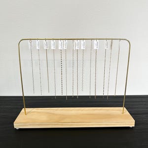 May include: A display stand with a light wood base and a gold-colored metal frame. The frame holds multiple silver necklaces, each with a different chain design. Small white tags with the word "INFINITE" are attached to each necklace.
