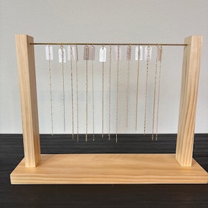 May include: A wooden jewelry display stand with a horizontal brass bar holding multiple gold necklaces. Each necklace hangs with a tag. The stand is made of light-colored wood and sits on a dark surface.