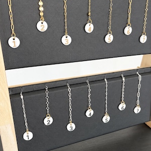 May include: A display stand holding fourteen necklaces arranged in two rows. The top row features seven gold-tone chains with white circular pendants numbered one through seven. The bottom row features seven silver-tone chains with white circular pendants numbered one through seven.