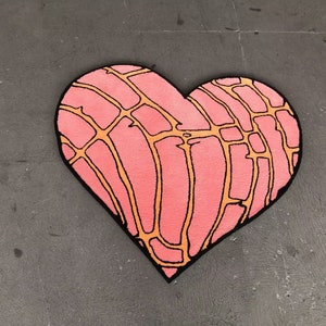 May include: A heart-shaped rug with a pink base and a grid-like pattern in orange, outlined in black. The rug is a soft floor covering, perfect for a bedroom or living room. The rug's design is reminiscent of a concha pastry.