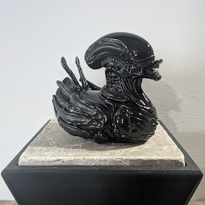 Può includere: Anatra di gomma nera a forma di Alien del franchise cinematografico. L'anatra ha una testa e un corpo dettagliati con una superficie testurizzata.