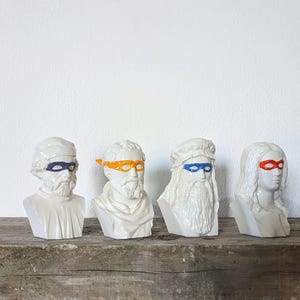 Puede incluir: Cinco bustos de cerámica blanca de figuras históricas famosas que llevan máscaras de superhéroes coloridas. Las máscaras son azules, naranja, azules, rojas y azules. Los bustos están dispuestos en fila sobre una superficie de madera.