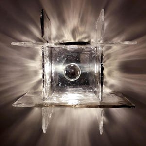 May include: A clear glass wall sconce with a square shape and a round light bulb in the centre. The sconce is made of multiple pieces of glass that are joined together.