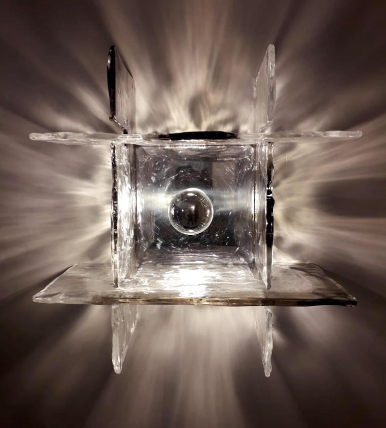 May include: A clear glass wall sconce with a square shape and a round light bulb in the centre. The sconce is made of multiple pieces of glass that are joined together.