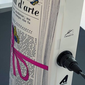 May include: A white ceramic lamp with a black cord and a black socket. The lamp features a printed design of a newspaper page with a pink ribbon and a butterfly. The text on the newspaper page reads "L. 1,500", "Fornasetti", and "mi d'arte".