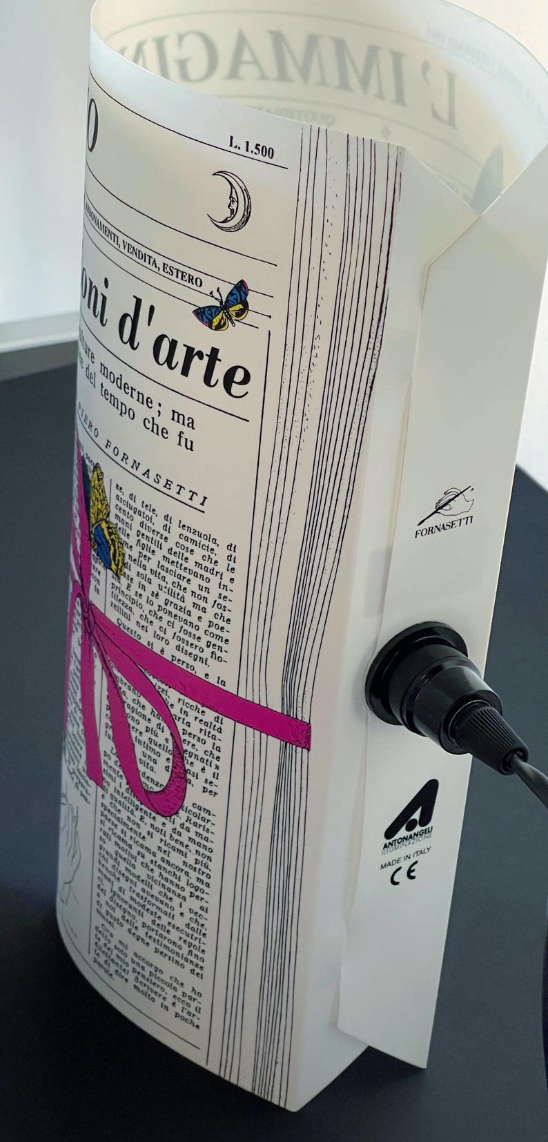 May include: A white ceramic lamp with a black cord and a black socket. The lamp features a printed design of a newspaper page with a pink ribbon and a butterfly. The text on the newspaper page reads "L. 1,500", "Fornasetti", and "mi d'arte".