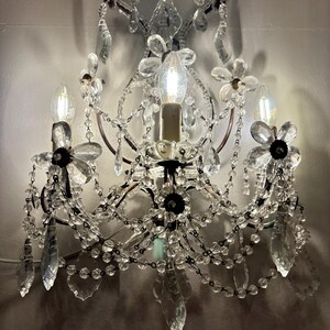 May include: A wall-mounted chandelier with clear crystal accents and flower-shaped details. The fixture has three light bulbs and decorative crystal droplets. The frame is a dark bronze color, and the overall design is ornate and elegant.