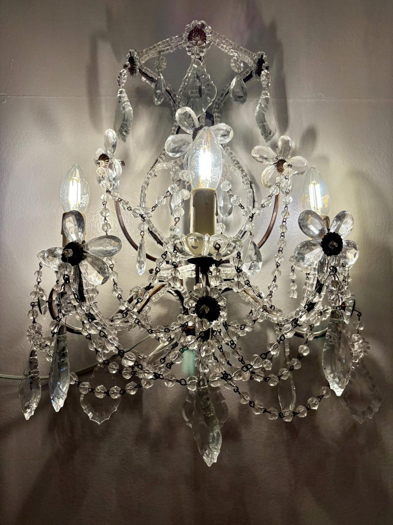 May include: A wall-mounted chandelier with clear crystal accents and flower-shaped details. The fixture has three light bulbs and decorative crystal droplets. The frame is a dark bronze color, and the overall design is ornate and elegant.