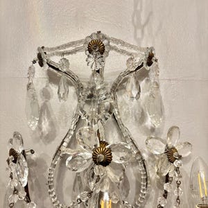 May include: An ornate wall sconce with a floral design. The sconce features clear crystal accents, including teardrop-shaped pendants and flower-shaped embellishments. The metal frame has a dark finish, and the sconce includes a light bulb.