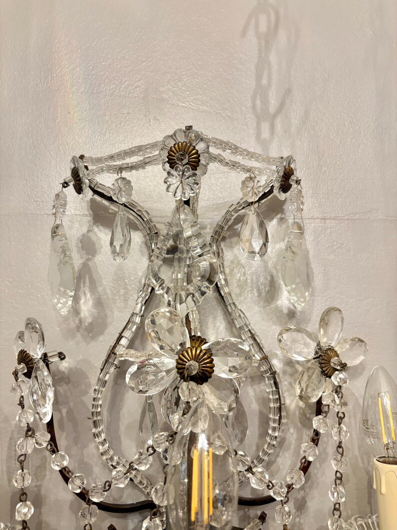 May include: An ornate wall sconce with a floral design. The sconce features clear crystal accents, including teardrop-shaped pendants and flower-shaped embellishments. The metal frame has a dark finish, and the sconce includes a light bulb.