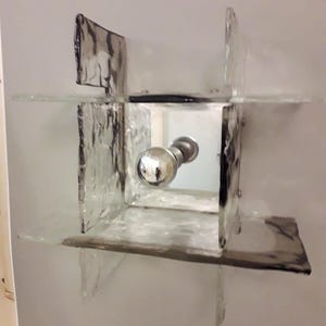 May include: A square-shaped wall sconce made of clear glass with a frosted texture. The sconce has a mirrored centre panel and a silver knob.