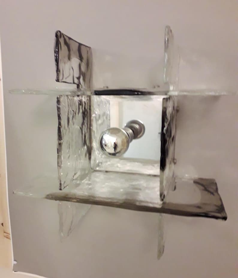 May include: A square-shaped wall sconce made of clear glass with a frosted texture. The sconce has a mirrored centre panel and a silver knob.