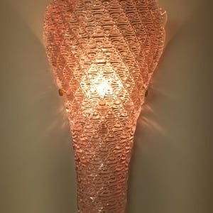May include: A pink glass wall sconce with a textured, bubble-like design. The sconce is lit and casts a warm glow.