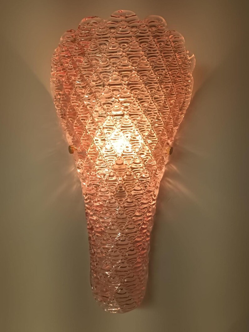 May include: A pink glass wall sconce with a textured, bubble-like design. The sconce is lit and casts a warm glow.