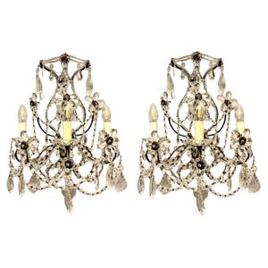 May include: Two ornate wall sconces with crystal and glass bead accents. Each sconce features three light fixtures, floral details, and hanging crystal pendants. The sconces have a black metal frame and are set against a white background.