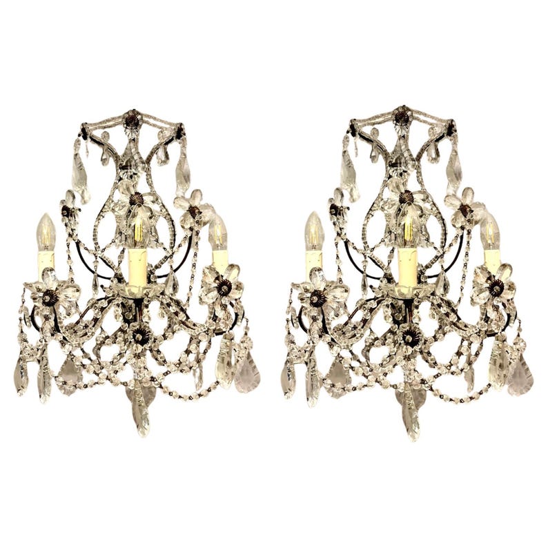 May include: Two ornate wall sconces with crystal and glass bead accents. Each sconce features three light fixtures, floral details, and hanging crystal pendants. The sconces have a black metal frame and are set against a white background.