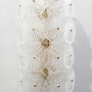 May include: A wall sconce featuring a vertical arrangement of clear, textured glass discs. The discs are arranged in a layered pattern and are attached to a gold-colored metal frame. Each disc has a small, gold-colored center.